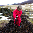 A child in a red 'Originals Waterproof Puddle Suit' stands on a pile of dark mulch, holding a mossy stick, with a field in the background.